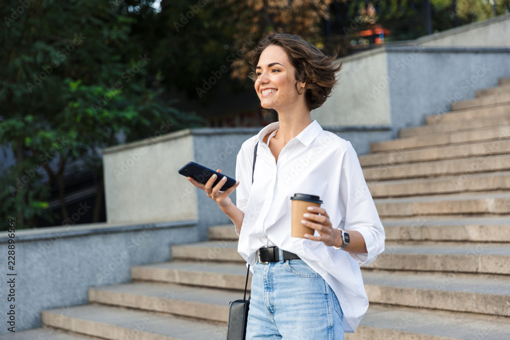 Cute beautiful woman walking on the street using mobile phone holding coffee.