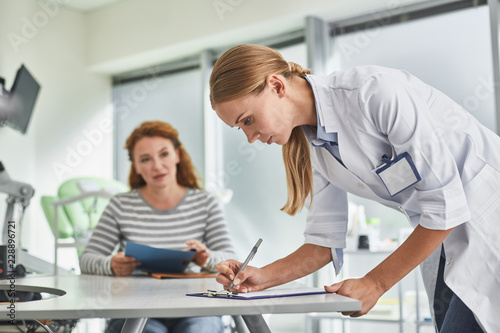 Paperwork. Portrait of gynecologist in white lab coat writing on clipboard while red-haired woman looking at her
