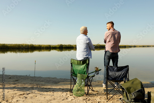 Father and adult son fishing together from riverside on sunny day