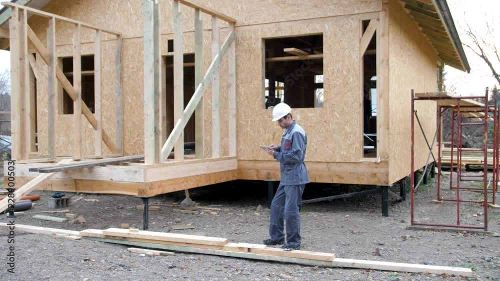 Caucasian worker in front of a house. Architect or builder check plans in a half built timber frame house. Builder on a construction site with a tablet. 4k Caucasian worker in front of a house. Architect or builder check plans in a half built timber frame house. Builder on a construction site with a tablet. 4k
