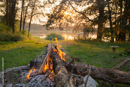 Lake side campfire with wood burning in a scenic setting during the sunset. Sun shines through canopy trees in golden light.