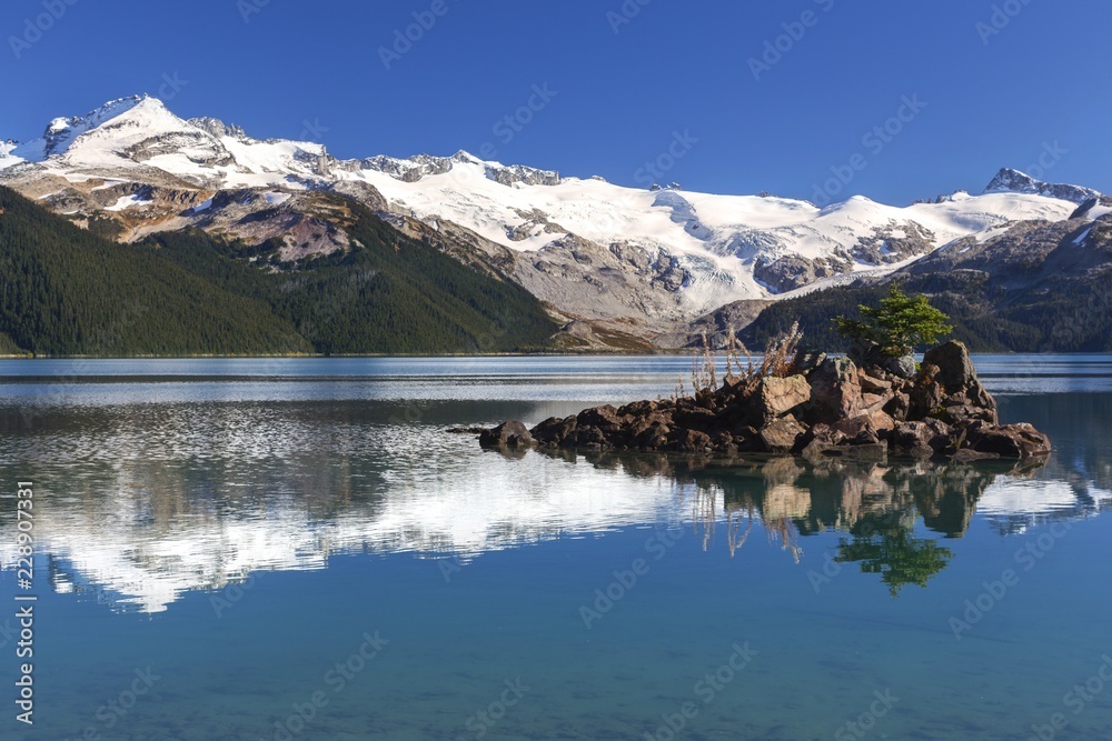 Isolated Pine Tree on a Rock Island and Distant Snowcapped Mountain Peaks Reflected in Calm Water of Garibaldi Lake in Coast Mountains of British Columbia Canada