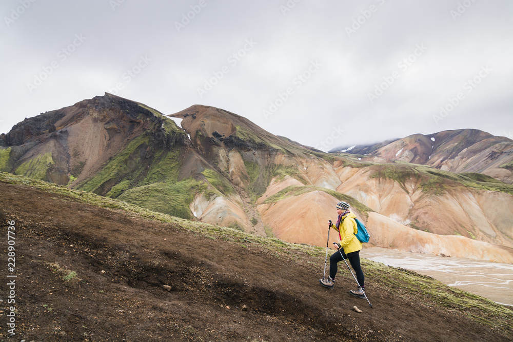 Woman in yellow raincoat hiking uphill in the colourful mountains in Landmannalaugar national park, Iceland