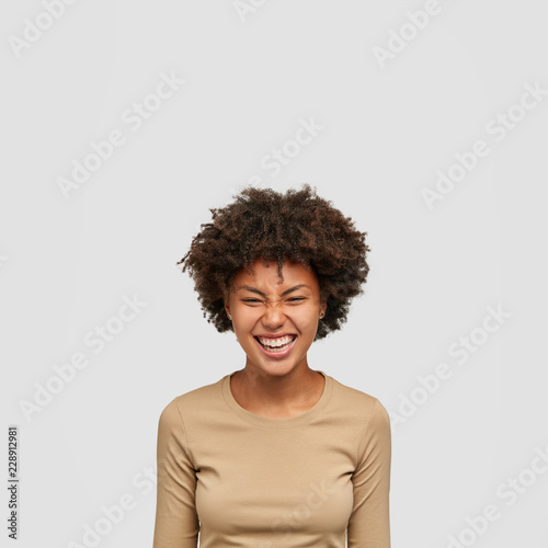 Indoor shot of happy woman has curly bushy hair, smiles broadly, squints face with happiness, has fun together with friends during weekned, dressed in comforable clothes, models in white studio © wayhome.studio 