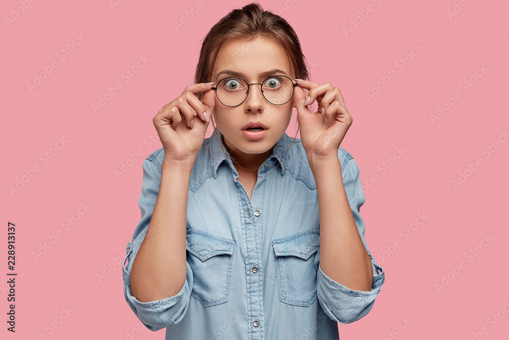 Emotive young Caucasian woman stares though big eyewear, hears something awesome and unbelievable, dressed in fashionable clothes, isolated over pink background. Emotions and reaction concept Emotive young Caucasian woman stares though big eyewear, hears something awesome and unbelievable, dressed in fashionable clothes, isolated over pink background. Emotions and reaction concept