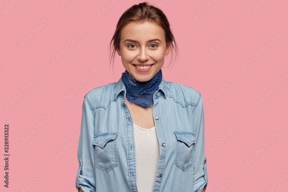 Φωτογραφία Pleasant looking lady with healthy skin, charming smile, wears bandana on neck, jean jacket, looks happily directly into camera, isolated over pink background