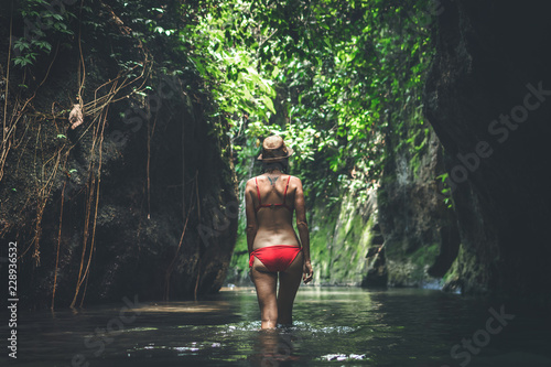 Rear view of young woman tourist with straw hat and red swimsuit in the deep jungle. Real adventure concept. Bali island.