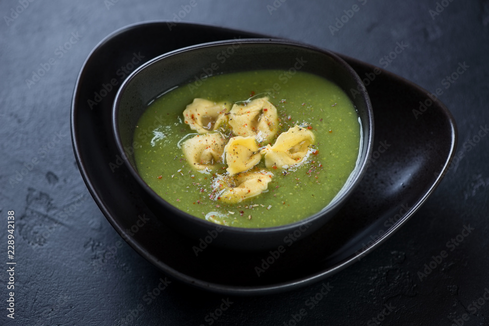Broccoli cream-soup with addition of tortellini served in a black tableware, horizontal shot over black stone background Broccoli cream-soup with addition of tortellini served in a black tableware, horizontal shot over black stone background