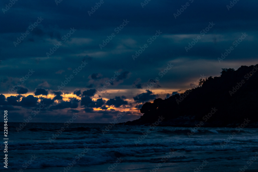 An amazing golden sunset behind a mountain silhouette edge on the end of Karon Beach, Phuket Thailand. A dramatic sky with vibrant light and reflections in the ocean