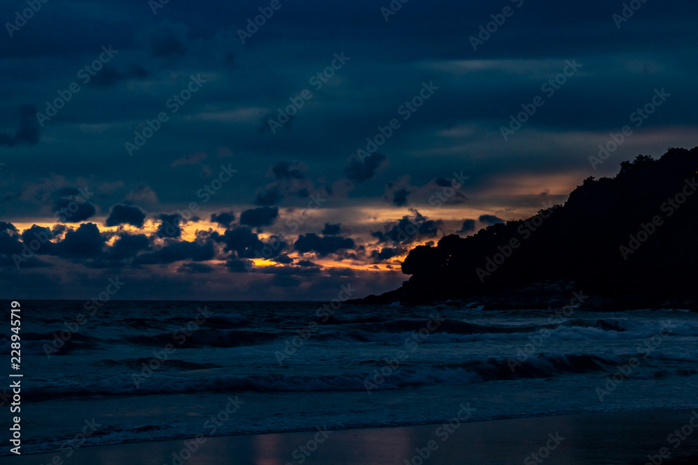 An amazing golden sunset behind a mountain silhouette edge on the end of Karon Beach, Phuket Thailand. A dramatic sky with vibrant light and reflections in the ocean