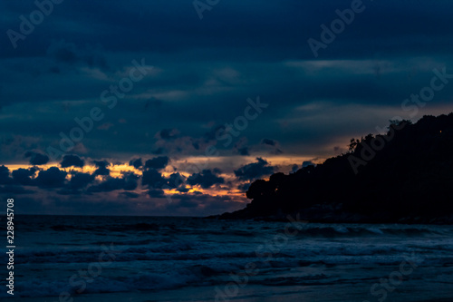 Obraz na płótnie An amazing golden sunset behind a mountain silhouette edge on the end of Karon Beach, Phuket Thailand. A dramatic sky with vibrant light and reflections in the ocean w salonie