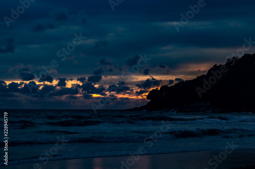 Obraz na płótnie An amazing golden sunset behind a mountain silhouette edge on the end of Karon Beach, Phuket Thailand. A dramatic sky with vibrant light and reflections in the ocean w salonie
