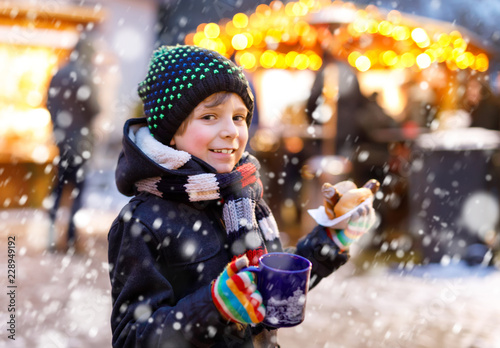 Little cute kid boy eating German sausage and drinking hot children punch on Christmas market. Happy child on traditional family market in Germany, Munich. Laughing boy in colorful winter clothes