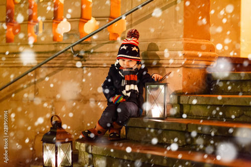Little cute kid boy with with a light lantern on stairs near church. Happy child on Christmas market in Germany. Kid waiting on parents on cold winter day