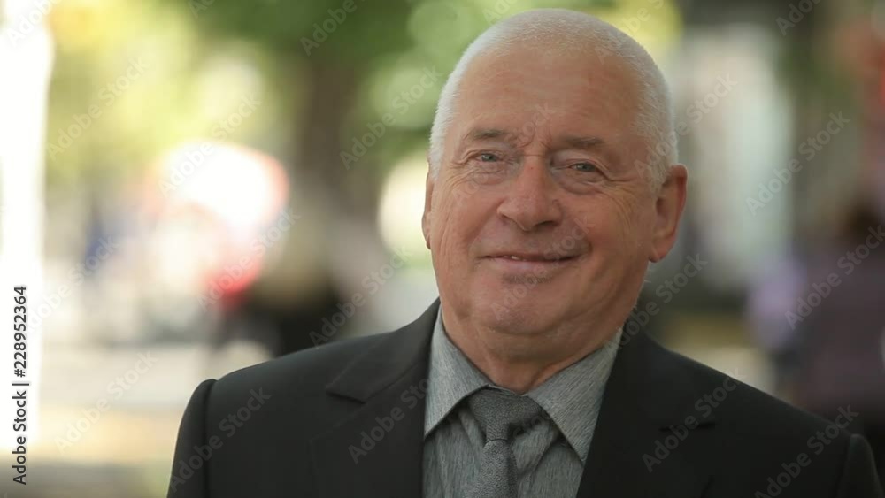 Cheerful bokeh portrait of an expirienced old man in a black business suit standing and smiling happily in a green street in autumn Cheerful bokeh portrait of an expirienced old man in a black business suit standing and smiling happily in a green street in autumn