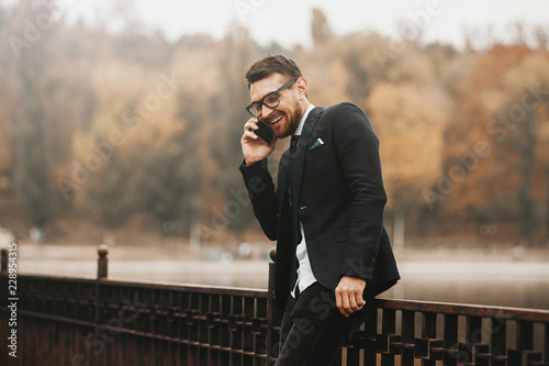 Cheerful young bearded man in suit talking on phone and smiling outdoor in park