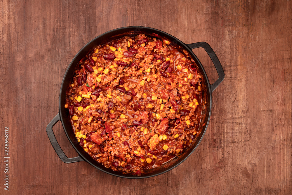 A large cast iron pan with chili con carne, shot from above on a dark rustic wooden table with a place for text