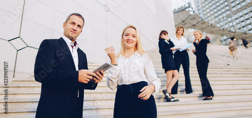 Fényképezés Boss keeping tablet and speaking with blonde secretary on stairs with biz partners in background