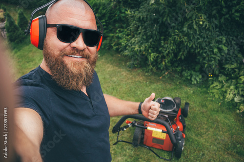 Slika na platnu Young bearded man making selfie while mows the lawn, cutting down the grass in t