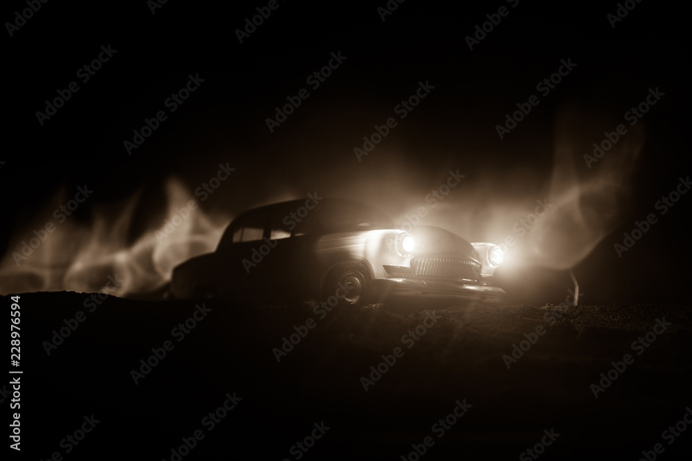 Silhouette of old vintage car in dark foggy toned background with glowing lights in low light.