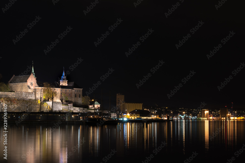 A beautiful night city. Lights of a night city on the coast. The historic castle at night. Reflections of lights on the water. Lights on the water. Calm sea at night. Oslo, Norway