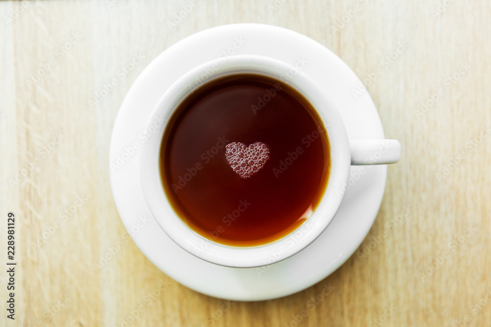 White cup of black tea with bubbles in a shape of heart. White cup on wooden table White cup of black tea with bubbles in a shape of heart. White cup on wooden table