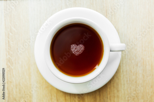 White cup of black tea with bubbles in a shape of heart. White cup on wooden table