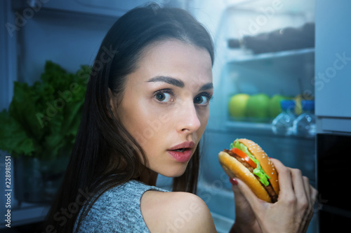 Hungry woman eating burger at night near fridge