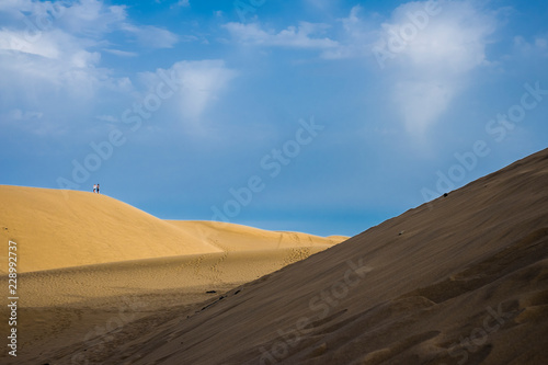 Couple of tourist standing on the hill of a dune in the yellow sandy desert. contrast of colors with blue celar sky. light and shadow. amazing natural outdoors background
