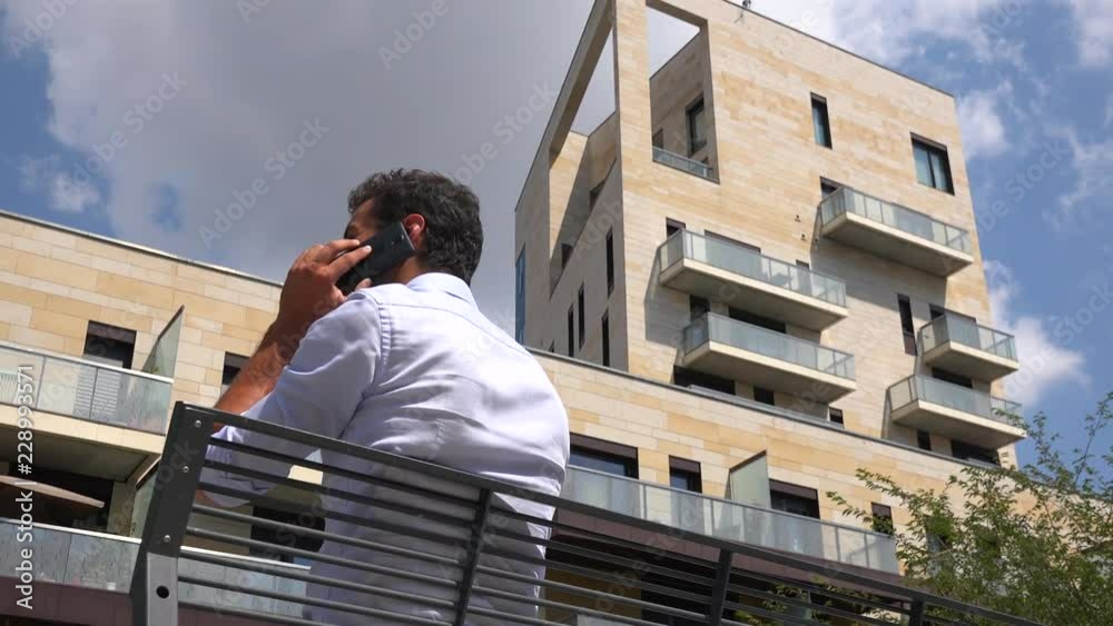 A businessman sits on a bench in front of an office building and talks on a smartphone - view from below A businessman sits on a bench in front of an office building and talks on a smartphone - view from below