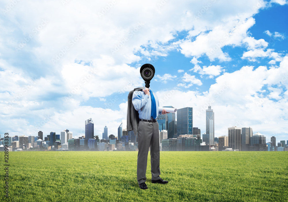 Camera headed man standing on green grass against modern cityscape Camera headed man standing on green grass against modern cityscape