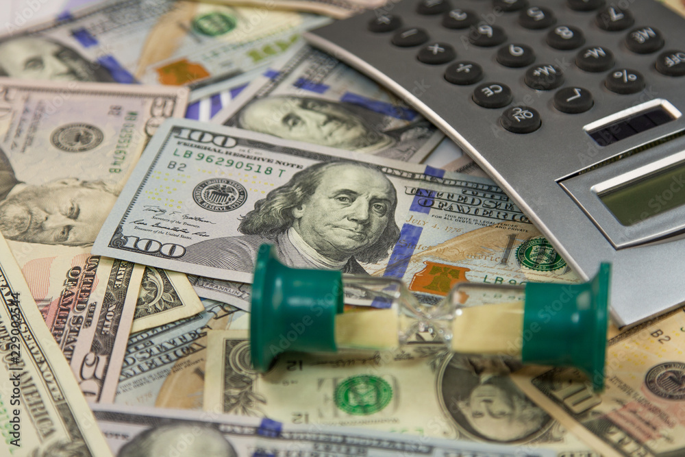 Fototapeta premium Hourglass is on dollar banknotes and calculator lies on a money background. Business and finance concept.