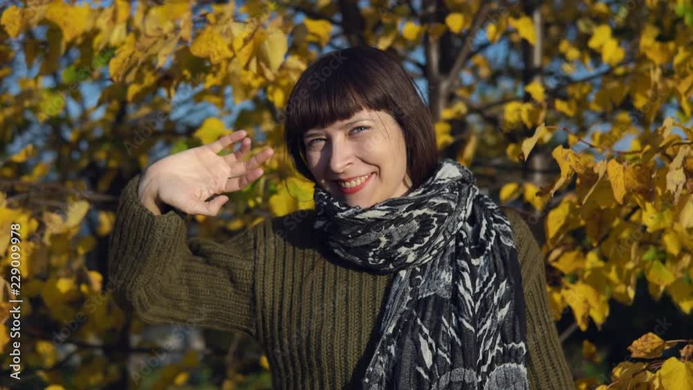 A young happy woman in a green knitted sweater waves her hand and greets someone against the yellow foliagein the city park in Indian summer.