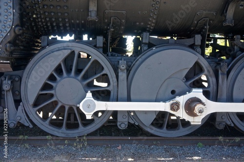 Close-up of the wheels of a locomotive; charcoal grey with bright metal pistons.