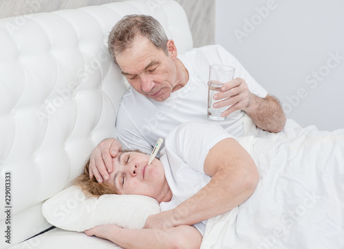 Senior man with glass of the water touches forehead his wife, checking the temperature