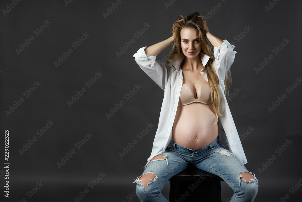 Beautiful smiling pregnant young blond woman, wearing ripped jeans, bra and  unbuttoned white shirt, sits and touches her long hair with her hand. Big  naked belly. Gray background Stock Photo | Adobe