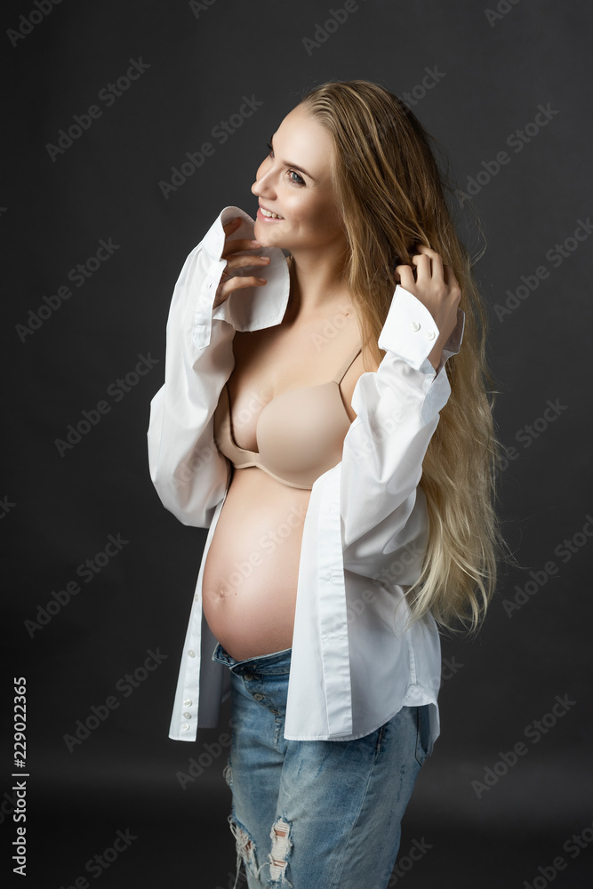 Beautiful smiling pregnant young blond woman, wearing ripped jeans, bra and  unbuttoned white shirt, stays and touches her face. Naked belly. Gray  background. Lifestyle, pregnancy, health Stock Photo | Adobe Stock