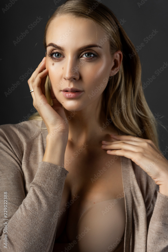 A beautiful blonde girl model wearing a knitted beige dress, unbuttoned on her breasts and a bra touches her face with her hand. Nude makeup. Clean, healthy skin. Gray background A beautiful blonde girl model wearing a knitted beige dress, unbuttoned on her breasts and a bra touches her face with her hand. Nude makeup. Clean, healthy skin. Gray background