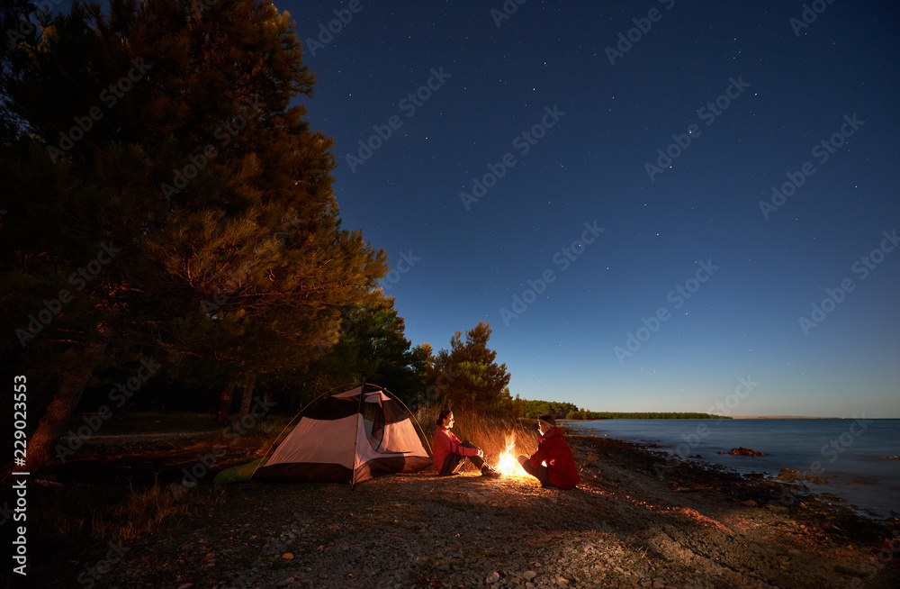 Night camping on sea shore. Happy young man and woman hikers relaxing in front of tent at campfire under evening sky on clear blue water and green forest background. Active lifestyle concept. - obrazy, fototapety, plakaty 