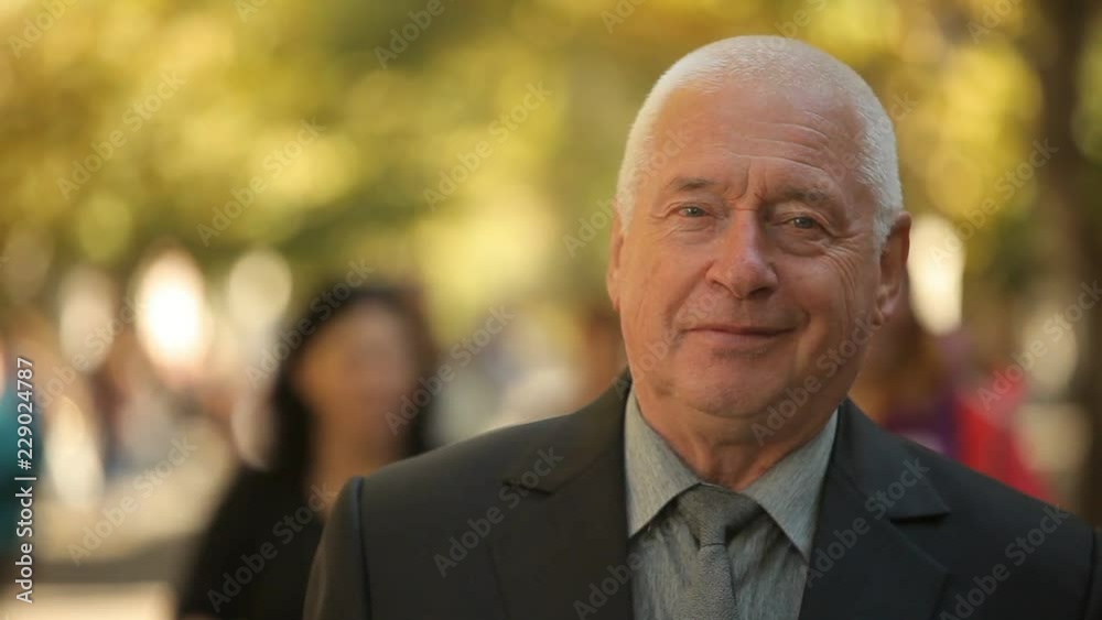 Cheerful view of a stylish white-haired man in a black business suit and a tie standing and smiling in a wonderful golden park in autumn Cheerful view of a stylish white-haired man in a black business suit and a tie standing and smiling in a wonderful golden park in autumn