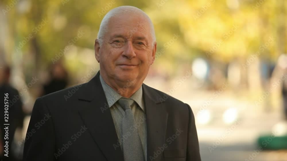 Exciting view of a wise senior man in a black business suit standing, smiling and enyoing life in a street with golden leafy trees in early autumn Exciting view of a wise senior man in a black business suit standing, smiling and enyoing life in a street with golden leafy trees in early autumn