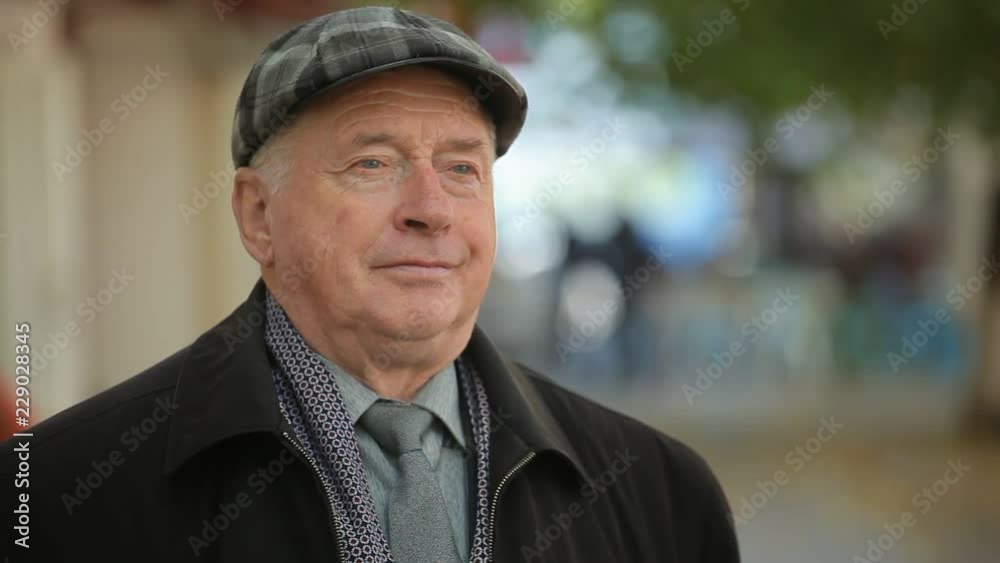 Amazing bokeh view of a neat white-haired man in a plaid flat cap standing and smiling cheerfully in a green street with people in autumn Amazing bokeh view of a neat white-haired man in a plaid flat cap standing and smiling cheerfully in a green street with people in autumn