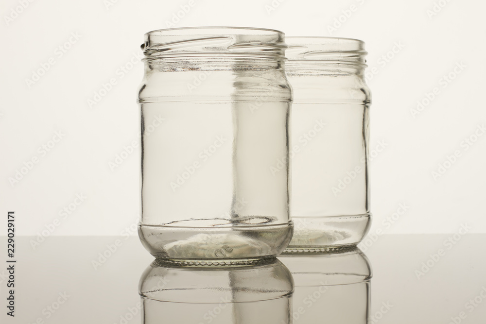 Two glass transparent empty jars. White background. Reflective surface. Two glass transparent empty jars. White background. Reflective surface.
