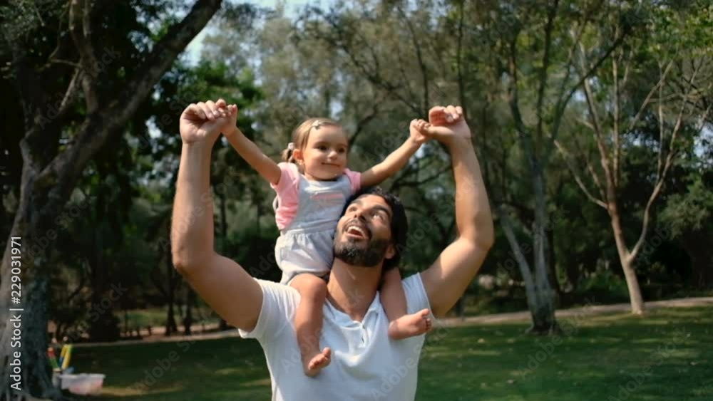 Caucasian father carrying on shoulders happy daughter in park. Smiling parent carrying little girl in summer park, side view. Childhood concept Caucasian father carrying on shoulders happy daughter in park. Smiling parent carrying little girl in summer park, side view. Childhood concept