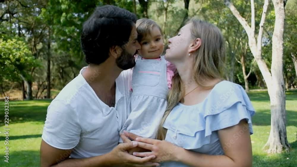 Cheerful parents hugging and kissing little daughter in sunny park, front view. Happy mother and father holding girl in slow motion. Happy family concept Cheerful parents hugging and kissing little daughter in sunny park, front view. Happy mother and father holding girl in slow motion. Happy family concept