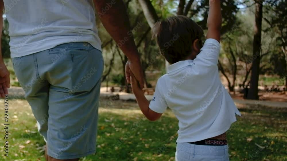 Little son pointing up while walking with father in park. Dad with boy holding hands and walking in park in slow motion. Family concept Little son pointing up while walking with father in park. Dad with boy holding hands and walking in park in slow motion. Family concept
