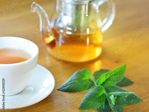White cup and glass teapot of herbal tea with fresh mint twig on wooden table