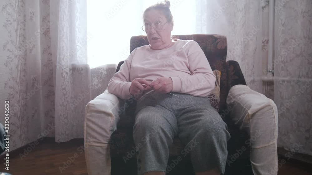 An elderly woman enjoys her hobby sitting in a chair and knitting on the background of the window. An elderly woman enjoys her hobby sitting in a chair and knitting on the background of the window.