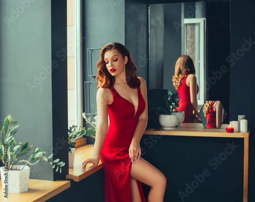 hot young adult, proud and domineering woman dressed in a long scarlet red long dress, sexually demonstrates her naked graceful leg, crouching on the window sill, room in loft stile with mirrors