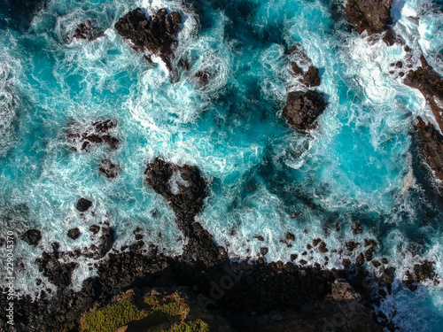 Aerial top view of sea waves hitting black volcanic rocks on the coastline with turquoise sea water. Amazing rock cliff seascape in the Portuguese coastline. Azores islands. Drone shot.